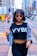 A stylish woman with short hair wearing round sunglasses, a black crop top with 'VYBE' printed on it, and a denim jacket draped over her shoulders. She stands confidently in an urban setting with blurred city buildings in the background.