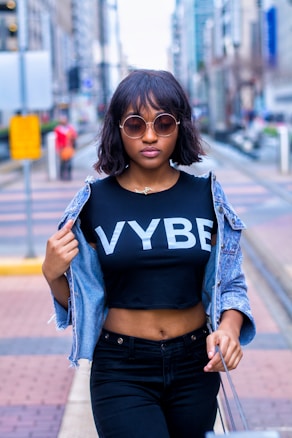 A stylish woman with short hair wearing round sunglasses, a black crop top with 'VYBE' printed on it, and a denim jacket draped over her shoulders. She stands confidently in an urban setting with blurred city buildings in the background.