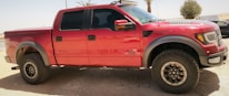 A red pickup truck with off-road tires and a rugged design is parked outdoors. The vehicle is equipped with black wheel arches, a black grille, and dark tinted windows. The background features a dry, desert-like setting with sparse vegetation, including palm trees and a bush.