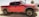 A red pickup truck with off-road tires and a rugged design is parked outdoors. The vehicle is equipped with black wheel arches, a black grille, and dark tinted windows. The background features a dry, desert-like setting with sparse vegetation, including palm trees and a bush.