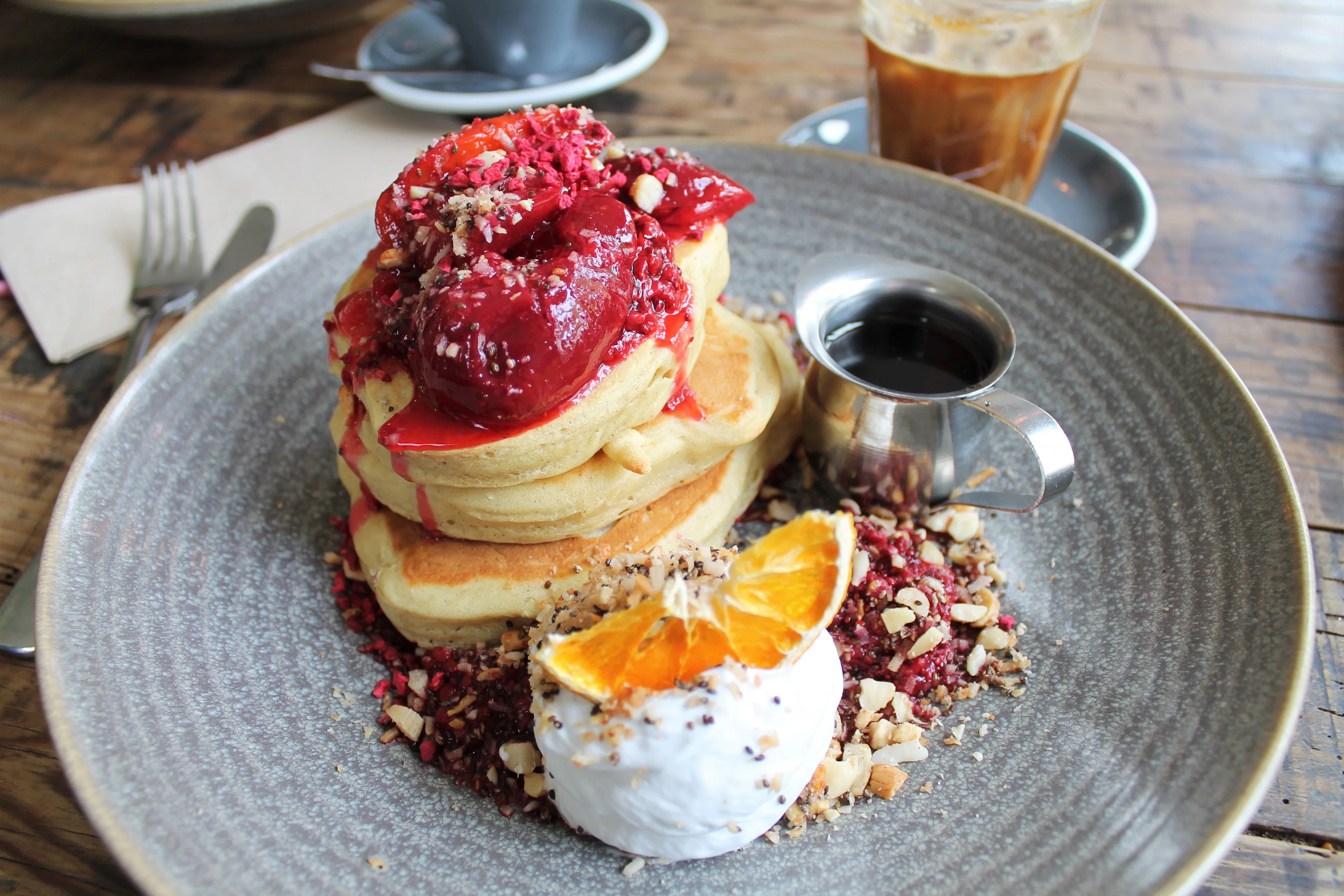 A stack of pancakes at a Queenstown cafe under a spoonful of plum compote and crushed nuts, with coconut yoghurt, a slice of orange, a small jug of syrup and an iced coffee on the scrubbed wooden table.
