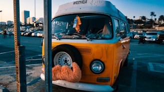 A vintage yellow Volkswagen van is parked in a lot with a spare tire mounted on the front, decorated with an orange furry cover. The background includes a parking lot filled with cars and tall buildings, along with palm trees indicating an urban setting. The sky is clear, suggesting a sunny day.