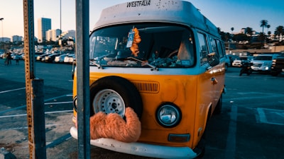 A vintage yellow Volkswagen van is parked in a lot with a spare tire mounted on the front, decorated with an orange furry cover. The background includes a parking lot filled with cars and tall buildings, along with palm trees indicating an urban setting. The sky is clear, suggesting a sunny day.
