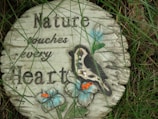 A round, decorative stone plaque placed on grass, featuring an engraved message and a colorful design. The message reads 'Nature touches every Heart.' The plaque includes the image of a bird perched on a branch with two large blue flowers and small orange details.