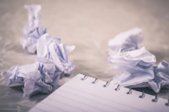 Crumpled sheets of paper on a wooden table beside a notepad