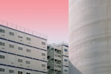 A modern architectural scene featuring a white modular building with multiple windows and a large adjacent cylindrical structure. The sky is a gradient of light pink and peach, creating a soft backdrop.