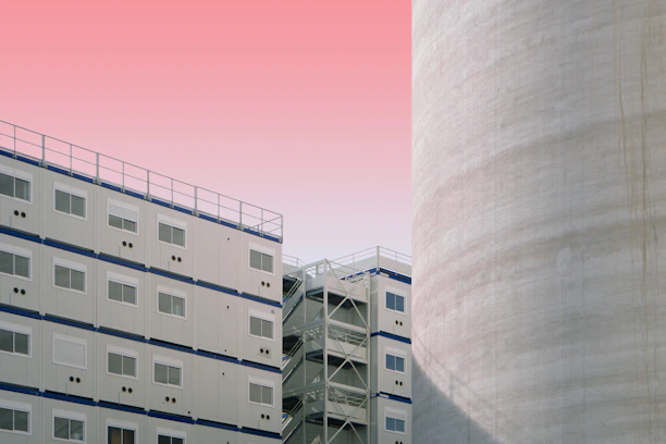 A modern architectural scene featuring a white modular building with multiple windows and a large adjacent cylindrical structure. The sky is a gradient of light pink and peach, creating a soft backdrop.