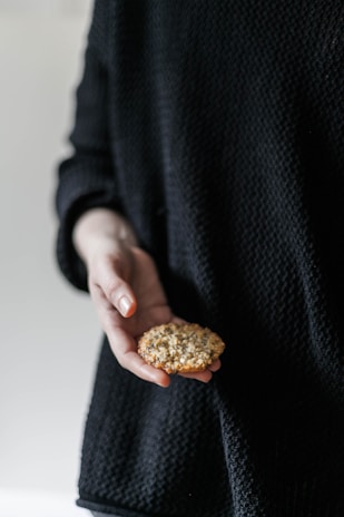 A person wearing a dark, textured sweater holds a cookie in their open hand. The cookie appears to be homemade with a rough, uneven texture, suggestive of a wholesome or artisanal bake.