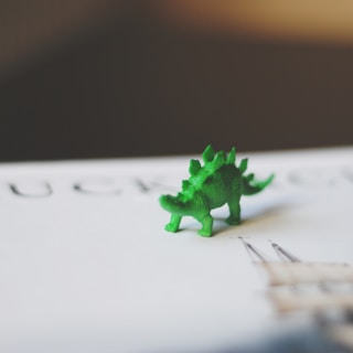 A small green toy dinosaur is placed on a surface with some blurred text and graphics in the background. The toy's texture and ridged back are clearly visible.