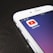 A smartphone screen displaying the YouTube app icon prominently. The phone is resting on a dark wooden surface, with the time and network information visible at the top.