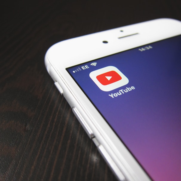 A smartphone screen displaying the YouTube app icon prominently. The phone is resting on a dark wooden surface, with the time and network information visible at the top.