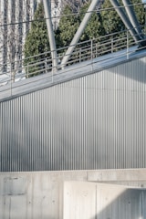 A modern architectural scene featuring a metal railing and geometric structures. The surface is composed of vertical corrugated metal panels, and there is a concrete wall in the foreground. In the background, green foliage and part of a glass building can be seen.
