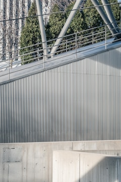 A modern architectural scene featuring a metal railing and geometric structures. The surface is composed of vertical corrugated metal panels, and there is a concrete wall in the foreground. In the background, green foliage and part of a glass building can be seen.