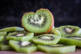 A close-up view of several slices of kiwi fruit, with one slice standing upright in the shape of a heart. The kiwi slices have a vibrant green color with small black seeds arranged in a circular pattern. The surface is visibly moist, suggesting freshness, and the background is dark, which enhances the vividness of the green fruit.