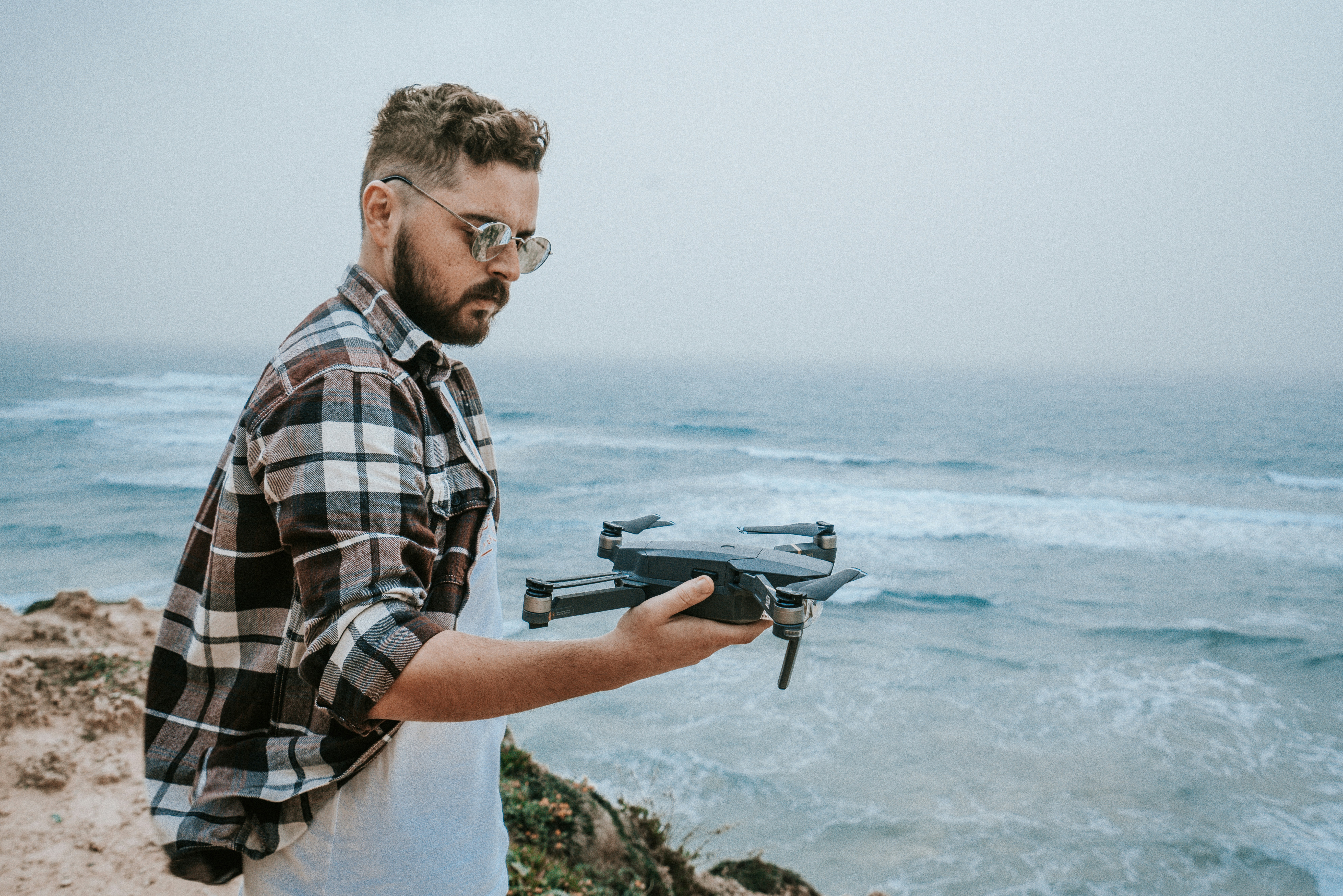 man holding black and brown DJI Mavic Pro drone on his right hand while standing on cliffside with view of sea during daytime photo, DJI’s Mavic Pro
