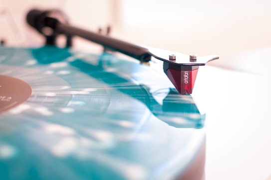 A close-up view of a vinyl record on a turntable with a stylus placed on it. The record is blue with light reflecting off its surface, creating a glossy effect. The stylus is red and detailed, signifying precision and care in design.