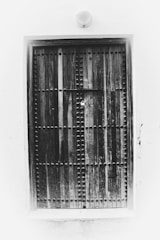 A large, rustic wooden door with a symmetrical pattern of metal studs set against a plain white wall. The door is locked with a padlock at the center, suggesting security or privacy. The overall texture appears weathered, adding an element of antiquity.