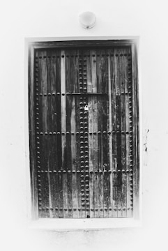 A large, rustic wooden door with a symmetrical pattern of metal studs set against a plain white wall. The door is locked with a padlock at the center, suggesting security or privacy. The overall texture appears weathered, adding an element of antiquity.
