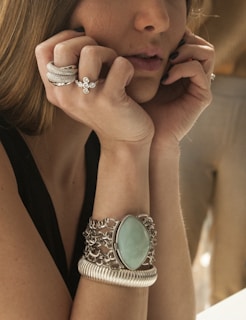 A woman is resting her face in her hands, showcasing multiple rings and bracelets on her fingers and wrists. The jewelry includes a prominent silver ring with a textured design, a diamond-studded ring, and a large bracelet with a turquoise stone set in an intricate silver chain design.