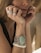 A woman is resting her face in her hands, showcasing multiple rings and bracelets on her fingers and wrists. The jewelry includes a prominent silver ring with a textured design, a diamond-studded ring, and a large bracelet with a turquoise stone set in an intricate silver chain design.