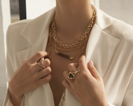 A close-up of a person wearing multiple gold necklaces and rings, featuring a white blazer. The jewelry includes chains and a statement ring, adding a touch of elegance and sophistication.