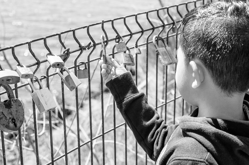 grayscale photography of boy holding padlock on fence