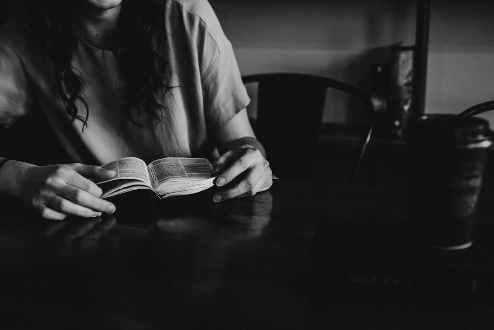 grayscale photo of woman reading a book on wooden table