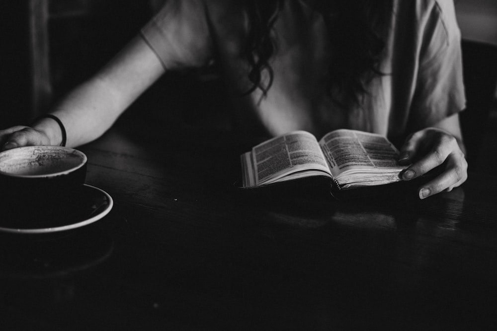 grayscale photo of woman reading book on table near black ceramic mug