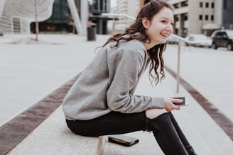 A young person with long hair, wearing a grey sweatshirt and ripped black jeans, is sitting on a concrete step. They are holding a smartphone and smiling. The background includes a modern urban environment with buildings, a road with a few cars, and some abstract structural elements.