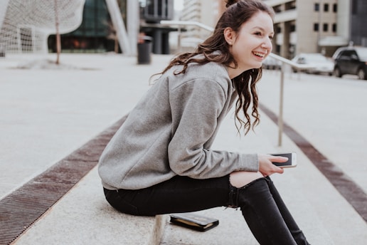 A young person with long hair, wearing a grey sweatshirt and ripped black jeans, is sitting on a concrete step. They are holding a smartphone and smiling. The background includes a modern urban environment with buildings, a road with a few cars, and some abstract structural elements.