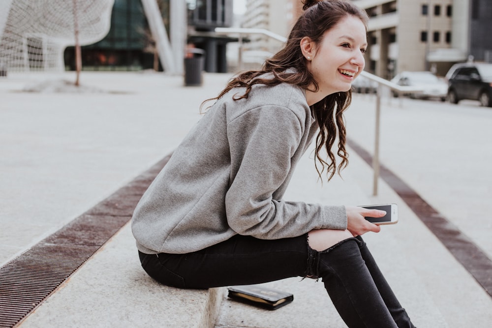 woman sits on sidewalk holding smartphone while smiling