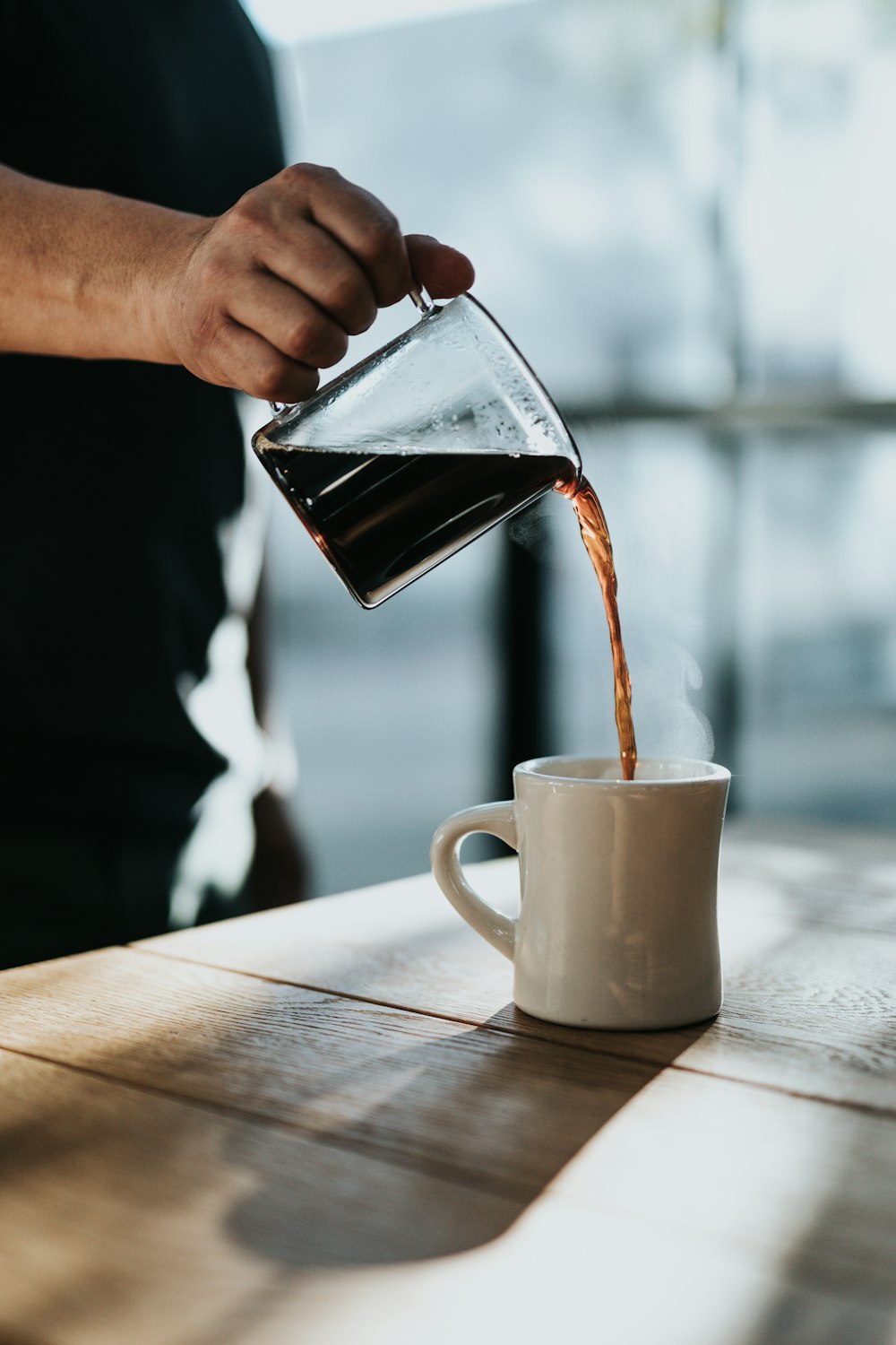 昼間、茶色の木製のテーブルに置かれた白い陶器のマグカップにブラックコーヒーを注ぐ人