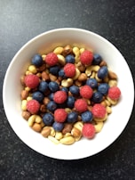 A white bowl filled with a mix of fruits and nuts. There are vibrant red raspberries and dark blue blueberries scattered on top of a variety of nuts, including almonds, cashews, and hazelnuts. The bowl is set against a dark textured background.