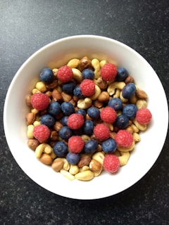 A white bowl filled with a mix of fruits and nuts. There are vibrant red raspberries and dark blue blueberries scattered on top of a variety of nuts, including almonds, cashews, and hazelnuts. The bowl is set against a dark textured background.