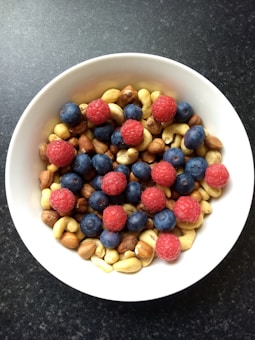 A white bowl filled with a mix of fruits and nuts. There are vibrant red raspberries and dark blue blueberries scattered on top of a variety of nuts, including almonds, cashews, and hazelnuts. The bowl is set against a dark textured background.
