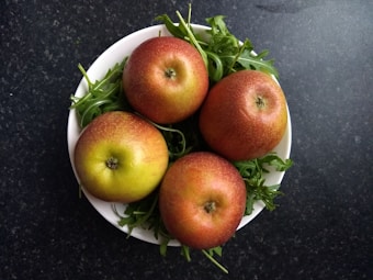 A white bowl contains four red apples placed on a bed of fresh green arugula. The bowl is set against a dark, speckled countertop surface.