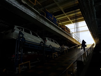 Several white cars are parked on an upper level of a multipurpose vehicle carrier. A person is walking up a ramp in a dimly lit industrial environment, with bright natural light visible at the far end.