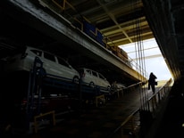 Several white cars are parked on an upper level of a multipurpose vehicle carrier. A person is walking up a ramp in a dimly lit industrial environment, with bright natural light visible at the far end.
