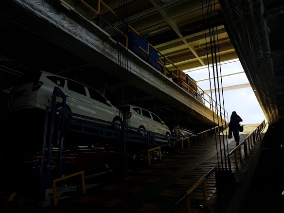 Several white cars are parked on an upper level of a multipurpose vehicle carrier. A person is walking up a ramp in a dimly lit industrial environment, with bright natural light visible at the far end.