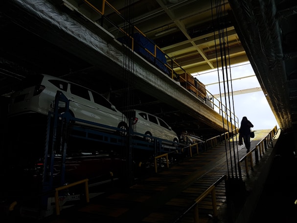 Several white cars are parked on an upper level of a multipurpose vehicle carrier. A person is walking up a ramp in a dimly lit industrial environment, with bright natural light visible at the far end.