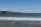 A beautiful coastal view with gentle waves lapping against the sandy shore. The horizon features rolling hills and scattered houses, with a clear sky overhead adorned with a few wispy clouds.