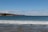 A beautiful coastal view with gentle waves lapping against the sandy shore. The horizon features rolling hills and scattered houses, with a clear sky overhead adorned with a few wispy clouds.