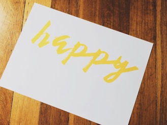 A piece of paper with the word 'happy' written in bold, yellow letters is placed on a wooden surface with a warm tone.