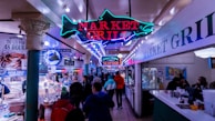 A bustling indoor seafood market featuring a prominent neon sign that reads 'Market Grill' with a fish design. The market has various stalls with fresh seafood on display, and several people are walking and shopping. The setting has an eclectic mix of bright lights, colorful signage, and a busy, vibrant atmosphere.
