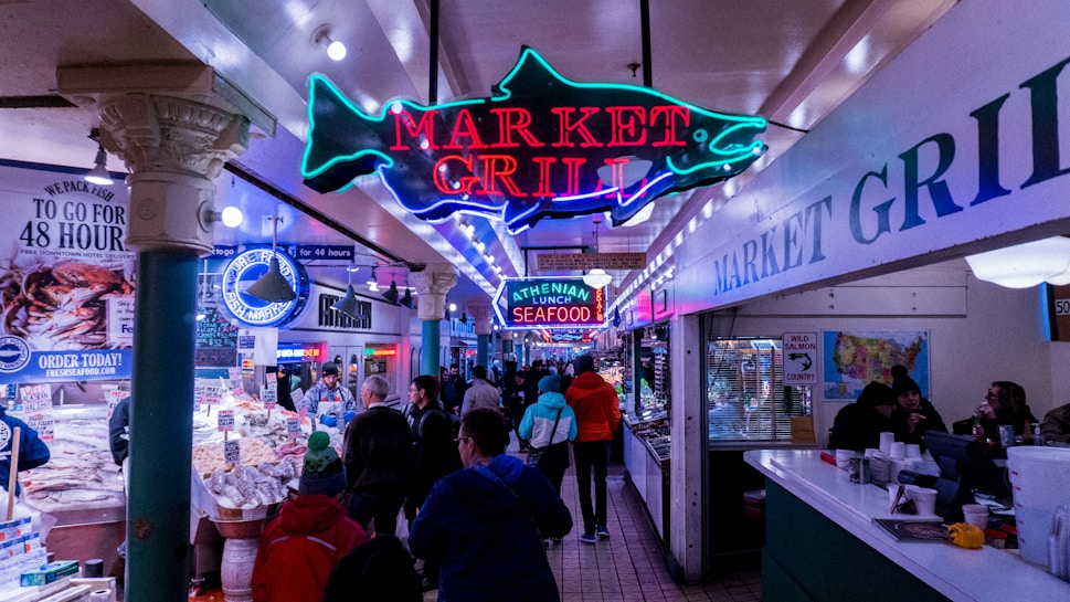 A bustling indoor seafood market featuring a prominent neon sign that reads 'Market Grill' with a fish design. The market has various stalls with fresh seafood on display, and several people are walking and shopping. The setting has an eclectic mix of bright lights, colorful signage, and a busy, vibrant atmosphere.