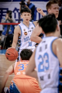 A basketball player in a white and blue jersey with the number 10 dribbles the ball on the court, surrounded by several other players. The focus is on the player with a determined expression, while the crowd and other players are slightly blurred, capturing the movement and intensity of the game.
