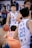 A basketball player in a white and blue jersey with the number 10 dribbles the ball on the court, surrounded by several other players. The focus is on the player with a determined expression, while the crowd and other players are slightly blurred, capturing the movement and intensity of the game.