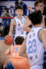 A basketball player in a white and blue jersey with the number 10 dribbles the ball on the court, surrounded by several other players. The focus is on the player with a determined expression, while the crowd and other players are slightly blurred, capturing the movement and intensity of the game.