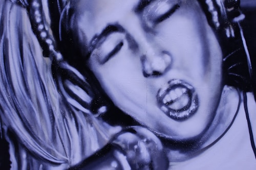 A monochromatic mural or graffiti depicts a person with long hair wearing headphones and passionately singing into a microphone. The image captures the intensity and emotion of a musical performance, highlighting the features and expressions with bold brushstrokes.