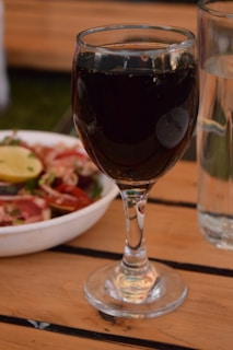 A glass of red wine is placed on a wooden table with a clear glass of water beside it. In the background, there is a white bowl filled with a fresh salad containing slices of lemon, tomatoes, and greens.